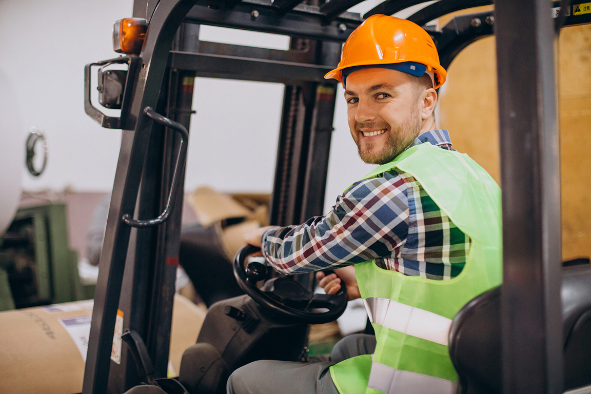 En leende förare i skyddshjälm och varselväst tittar sig om över axeln från sin plats på en gaffeltruck.