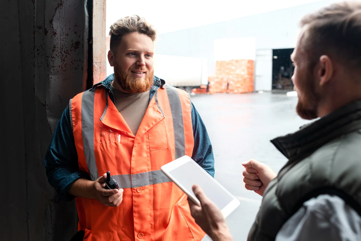 En medarbetare i varselväst pratar med en kollega vid en lastport, med en lastbil och pallgods i bakgrunden.