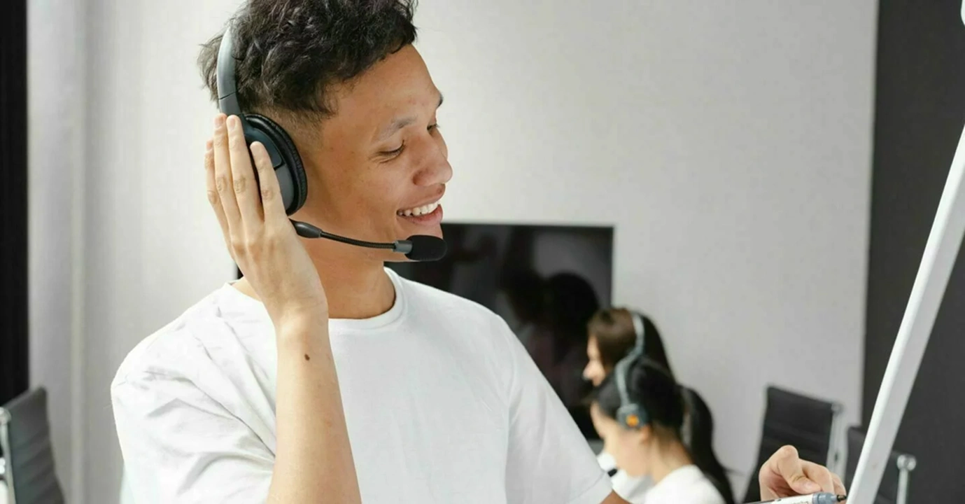 Kundtjänstmedarbetare med headset ler under ett samtal, med kollegor i bakgrunden.