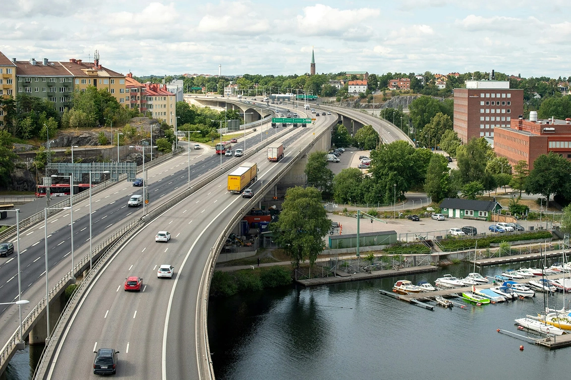 Vy över en flerfilig motorväg med trafik och en DHL-lastbil, med en småbåtshamn och stadsbebyggelse i bakgrunden.