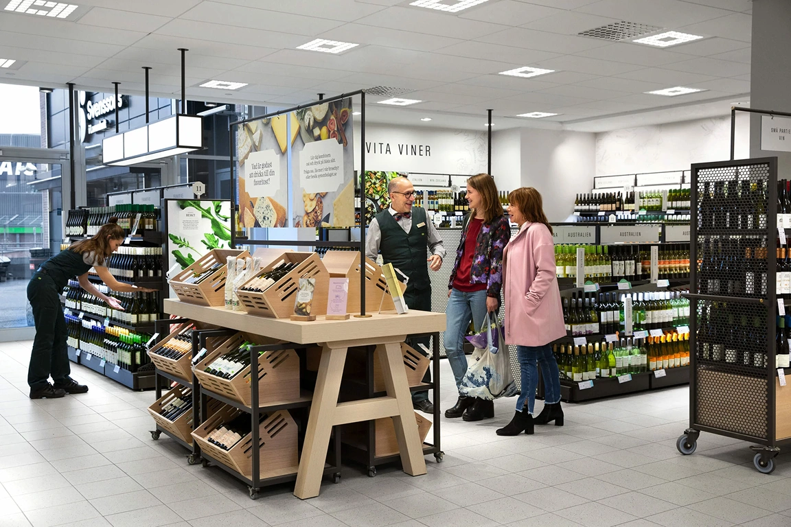 Butiksbiträde pratar med två kunder vid en vinavdelning med provbord och hyllor fulla av flaskor.