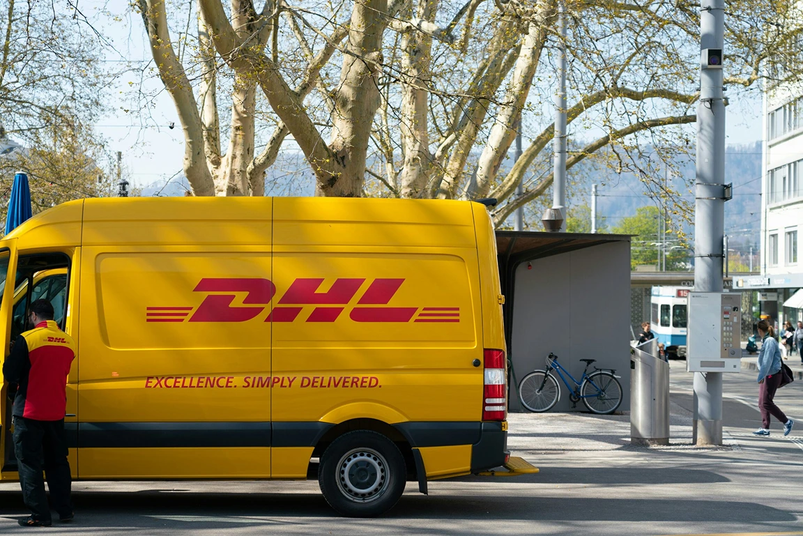 En gul DHL-skåpbil parkerad på en stadsgata, med en anställd i uniform vid föraren och en spårvagn i bakgrunden.
