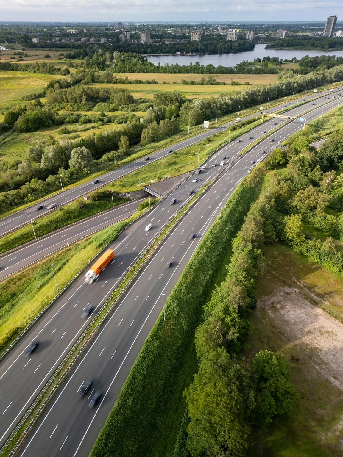 Flygfoto över en flerfilig motorväg i Sverige med bilar och en lastbil i rörelse, omgiven av gröna fält, skog och en sjö med en stad i bakgrunden.