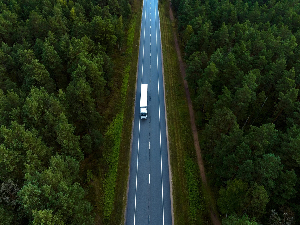 Flygfoto rakt uppifrån över en väg genom tät skog, med en vit lastbil på vägen.