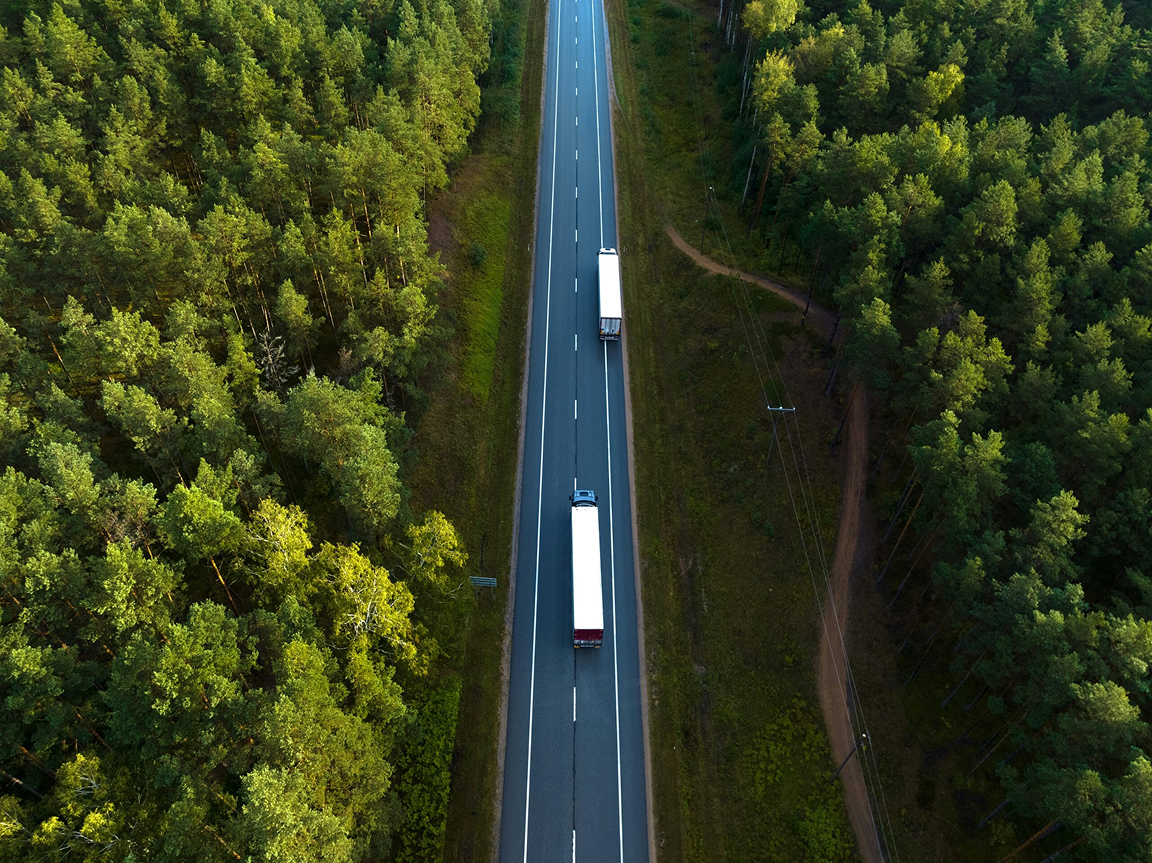 Flygfoto rakt uppifrån över en väg genom tät skog, med två vita lastbilar på väg i samma riktning.